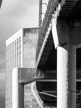 A black and white photograph captures a modern urban scene with a prominent high-rise building featuring numerous windows and reflective glass. In the foreground, there is a large concrete structure under construction, with visible beams and scaffolding. The contrast between the finished skyscraper and the developing construction creates a dynamic architectural composition.