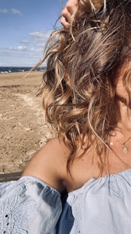 Blonde, wavy hair partially obscures the view of a sandy beach and ocean waves. A light blue, off-the-shoulder top is worn by the person, who has a necklace with a small pendant. The sky is mostly clear with some clouds, giving the scene a bright and sunny ambiance.