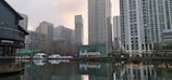 A modern urban waterfront setting with a series of tall buildings reflecting in the water. The structures are primarily composed of glass and steel, indicative of contemporary architecture. Several boats are docked alongside the water, and there is visible signage on a building with the word 'ballymore' in lowercase letters.