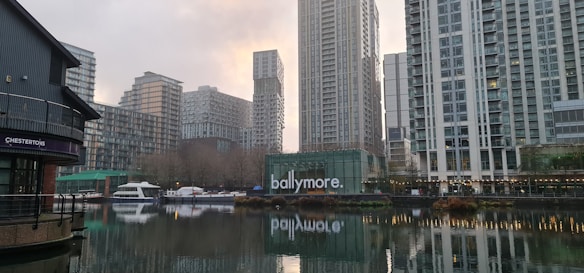 A modern urban waterfront setting with a series of tall buildings reflecting in the water. The structures are primarily composed of glass and steel, indicative of contemporary architecture. Several boats are docked alongside the water, and there is visible signage on a building with the word 'ballymore' in lowercase letters.