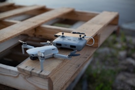 A drone and its remote controller resting on a wooden pallet near a body of water. The drone is compact with four propellers, and the remote controller has a smartphone attached to it.
