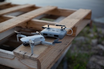 A drone and its remote controller resting on a wooden pallet near a body of water. The drone is compact with four propellers, and the remote controller has a smartphone attached to it.