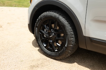 A close-up view of a car's black tire and rim, set against a sandy or gravel background. The vehicle's body is partially visible, showing a white or light gray exterior.