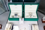 Two luxury watches displayed in elegant green boxes, each marked with the brand's logo. These watches are placed on a soft beige surface within a glass display case, surrounded by other jewelry items including bracelets and watches.