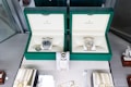 Two luxury watches displayed in elegant green boxes, each marked with the brand's logo. These watches are placed on a soft beige surface within a glass display case, surrounded by other jewelry items including bracelets and watches.