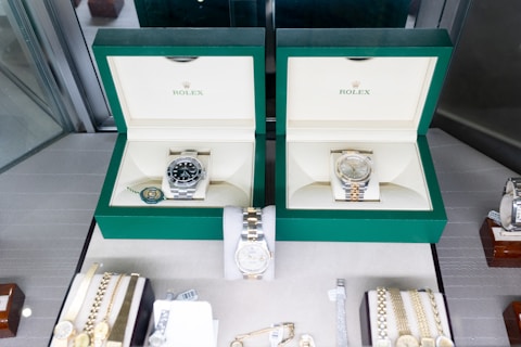 Two luxury watches displayed in elegant green boxes, each marked with the brand's logo. These watches are placed on a soft beige surface within a glass display case, surrounded by other jewelry items including bracelets and watches.