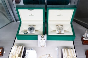 Two luxury watches displayed in elegant green boxes, each marked with the brand's logo. These watches are placed on a soft beige surface within a glass display case, surrounded by other jewelry items including bracelets and watches.