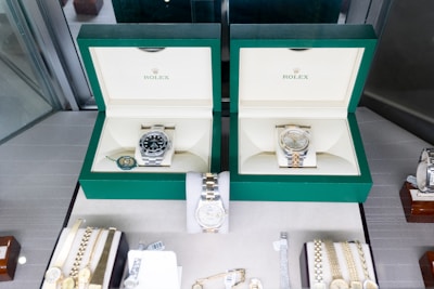 Two luxury watches displayed in elegant green boxes, each marked with the brand's logo. These watches are placed on a soft beige surface within a glass display case, surrounded by other jewelry items including bracelets and watches.