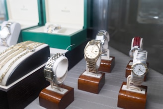 Several luxury watches are displayed on wooden stands, with some showcased in open boxes. The assortment includes different designs and metal finishes, including gold and stainless steel. Bracelets and watchbands are also visible, adding to the luxurious appeal.