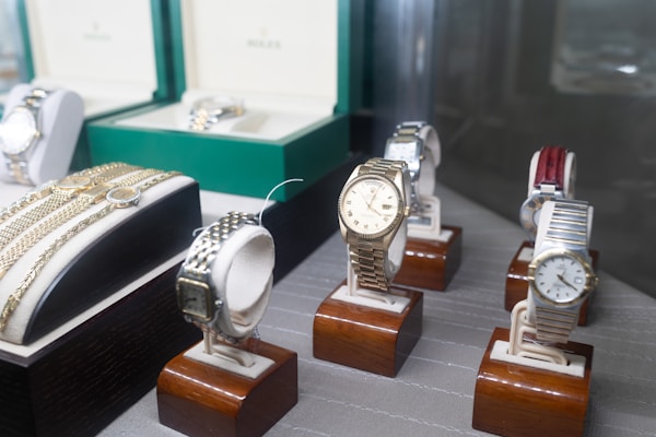 Several luxury watches are displayed on wooden stands, with some showcased in open boxes. The assortment includes different designs and metal finishes, including gold and stainless steel. Bracelets and watchbands are also visible, adding to the luxurious appeal.