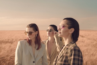 Three women wearing stylish suits and sunglasses stand in a vast golden wheat field under a clear sky. Their hair is sleek and brushed back, and they seem to be gazing into the distance, capturing a sense of elegance and poise.