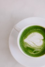 A white ceramic cup filled with green matcha latte, featuring a heart-shaped foam art in the center, placed on a white saucer against a light background.