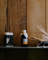 A wooden background provides a rustic setting for a shelf featuring a block of soap and a bottle wrapped with a rustic cap. The items are accompanied by a bowl filled with aromatic sticks, conveying a natural and soothing ambiance.