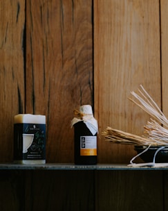 A wooden background provides a rustic setting for a shelf featuring a block of soap and a bottle wrapped with a rustic cap. The items are accompanied by a bowl filled with aromatic sticks, conveying a natural and soothing ambiance.