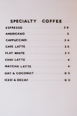 A neatly organized coffee menu on a white background, displaying various specialty coffee options such as espresso, americano, cappuccino, cafe latte, flat white, chai latte, matcha latte, oat and coconut, and iced and decaf, each followed by corresponding prices.