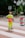 A construction worker wearing a bright neon yellow safety uniform and an orange cap stands on a paved area near a construction site. Behind them, fencing separates the pedestrian area from construction materials and equipment. Mature trees provide a leafy backdrop, and some urban buildings are visible in the distance.