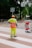 A construction worker wearing a bright neon yellow safety uniform and an orange cap stands on a paved area near a construction site. Behind them, fencing separates the pedestrian area from construction materials and equipment. Mature trees provide a leafy backdrop, and some urban buildings are visible in the distance.
