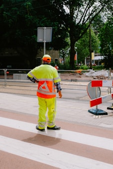A construction worker wearing a bright neon yellow safety uniform and an orange cap stands on a paved area near a construction site. Behind them, fencing separates the pedestrian area from construction materials and equipment. Mature trees provide a leafy backdrop, and some urban buildings are visible in the distance.