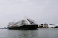 A large cruise ship is sailing on a calm body of water with several buildings and cranes visible in the background. The ship is adorned with multiple decks and features lifeboats and passenger windows along its side.