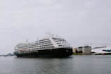 A large cruise ship is sailing on a calm body of water with several buildings and cranes visible in the background. The ship is adorned with multiple decks and features lifeboats and passenger windows along its side.