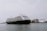 A large cruise ship is sailing on a calm body of water with several buildings and cranes visible in the background. The ship is adorned with multiple decks and features lifeboats and passenger windows along its side.