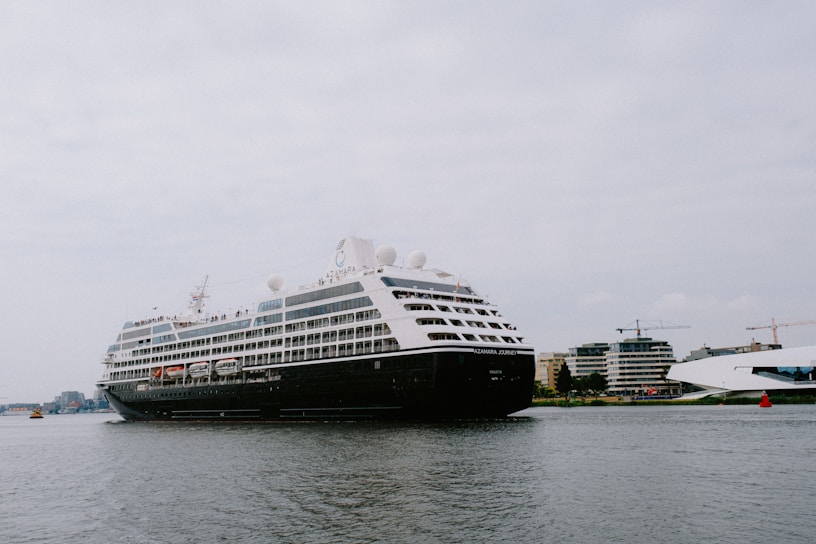 A large cruise ship is sailing on a calm body of water with several buildings and cranes visible in the background. The ship is adorned with multiple decks and features lifeboats and passenger windows along its side.