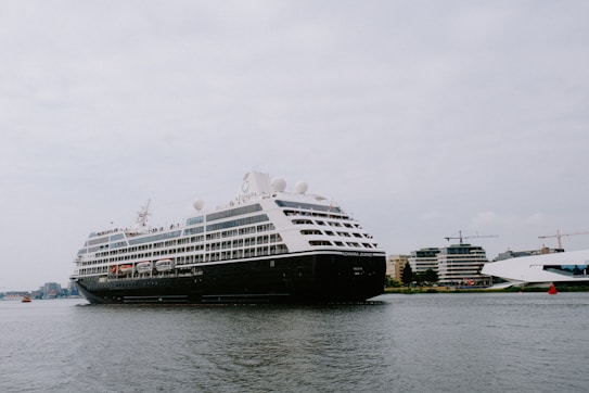 A large cruise ship is sailing on a calm body of water with several buildings and cranes visible in the background. The ship is adorned with multiple decks and features lifeboats and passenger windows along its side.