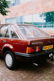 A vintage car with a brown exterior is parked on a brick pavement. The building in the background has large glass windows and stone tiles. The car has a visible license plate and an emblem that reads 'Escort 1.6 GL'.