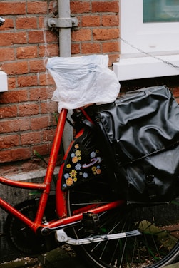 A red bicycle is parked against a brick wall. The seat is covered with a plastic bag, and there is a black bag attached to the back. The wall features a narrow, white-framed window, and there's a chain attached to the bike for security. Decorative flower patterns adorn the bicycle's frame.