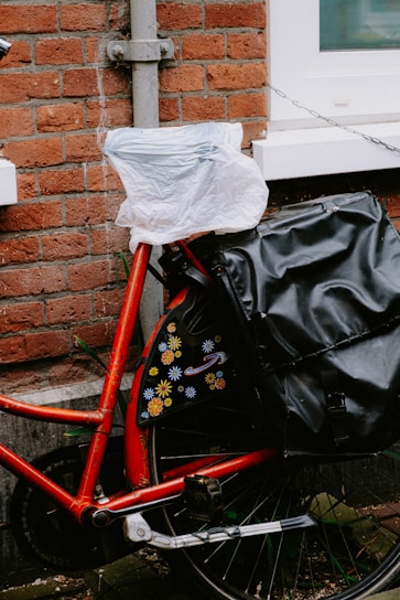 A red bicycle is parked against a brick wall. The seat is covered with a plastic bag, and there is a black bag attached to the back. The wall features a narrow, white-framed window, and there's a chain attached to the bike for security. Decorative flower patterns adorn the bicycle's frame.