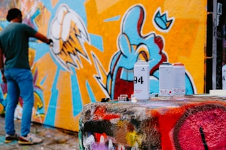 A person is spray-painting a colorful mural on a wall, which features vibrant designs with abstract shapes and sharp lines. In the foreground, several spray paint cans are placed on a surface covered with multicolored graffiti.