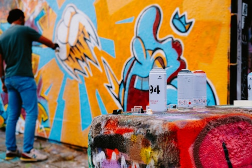 A person is spray-painting a colorful mural on a wall, which features vibrant designs with abstract shapes and sharp lines. In the foreground, several spray paint cans are placed on a surface covered with multicolored graffiti.