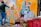 A person is spray-painting a colorful mural on a wall, which features vibrant designs with abstract shapes and sharp lines. In the foreground, several spray paint cans are placed on a surface covered with multicolored graffiti.