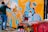 A person is spray-painting a colorful mural on a wall, which features vibrant designs with abstract shapes and sharp lines. In the foreground, several spray paint cans are placed on a surface covered with multicolored graffiti.