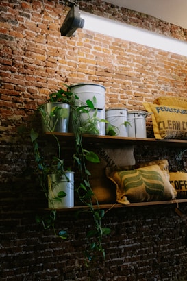 A rustic brick wall serves as the backdrop for a wooden shelf adorned with several metal buckets. Green plants with trailing vines are interspersed among these buckets. A few burlap sacks, featuring printed text, add to the decor, contributing to a cozy, vintage atmosphere.