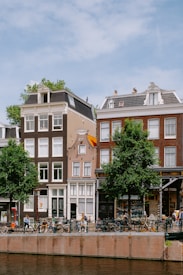 A picturesque canal-side street with traditional Dutch townhouses, featuring large windows and ornate details. Bicycles are parked along the street and several people are walking by. Trees line the street, and a calm canal runs parallel to the scene under a partly cloudy sky.