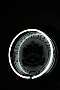 A dimly lit circular sign displaying the Delta Sigma Phi emblem, surrounded by a glowing neon ring. The emblem includes a shield with a variety of symbols, and text that reads 'EST 1899'. The background is predominantly dark, highlighting the neon effect and the emblem.