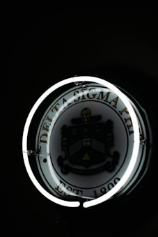 A dimly lit circular sign displaying the Delta Sigma Phi emblem, surrounded by a glowing neon ring. The emblem includes a shield with a variety of symbols, and text that reads 'EST 1899'. The background is predominantly dark, highlighting the neon effect and the emblem.
