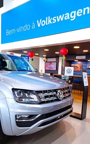 A Volkswagen showroom featuring a silver SUV prominently displayed in the foreground. The background includes a welcoming sign in Portuguese, 'Bem-vindo à Volkswagen,' and various decorations such as red lanterns. The space is well-lit with modern furnishings, and there is a cafe section visible.