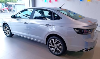 A sleek silver sedan is displayed in a well-lit showroom. The car has a modern design with aerodynamic lines and stylish alloy wheels. The showroom is decorated with colorful triangular flags hanging from the ceiling, adding a festive touch.