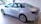 A sleek silver sedan is displayed in a well-lit showroom. The car has a modern design with aerodynamic lines and stylish alloy wheels. The showroom is decorated with colorful triangular flags hanging from the ceiling, adding a festive touch.