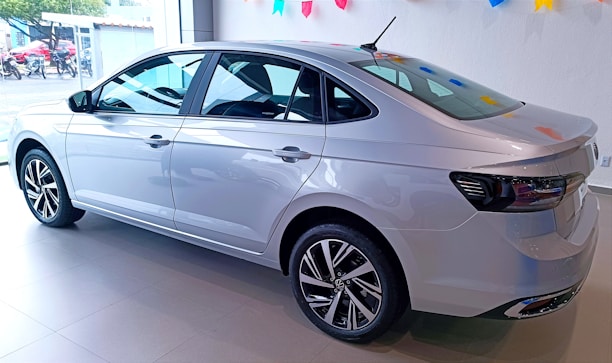 A sleek silver sedan is displayed in a well-lit showroom. The car has a modern design with aerodynamic lines and stylish alloy wheels. The showroom is decorated with colorful triangular flags hanging from the ceiling, adding a festive touch.