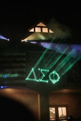 A building at night illuminated by neon green laser lights forming Greek letters on the facade. Smoke or fog enhances the atmospheric effect, and a portion of an American flag is visible on the building's wall. Warm light emanates from a window under a triangular roof.