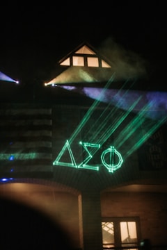 A building at night illuminated by neon green laser lights forming Greek letters on the facade. Smoke or fog enhances the atmospheric effect, and a portion of an American flag is visible on the building's wall. Warm light emanates from a window under a triangular roof.