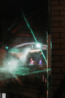 A dimly lit scene with a DJ performing on a balcony under colorful lights and smoke effects. The setting has a moody atmosphere, with beams of green and red light cutting through the mist. The surroundings are urban, with brick architecture visible.