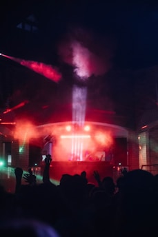 A vibrant club scene with colorful lighting effects, featuring a crowd of people with raised hands silhouetted against the bright lights. Smoky red and blue hues dominate the atmosphere as beams of light create a dynamic and energetic environment.