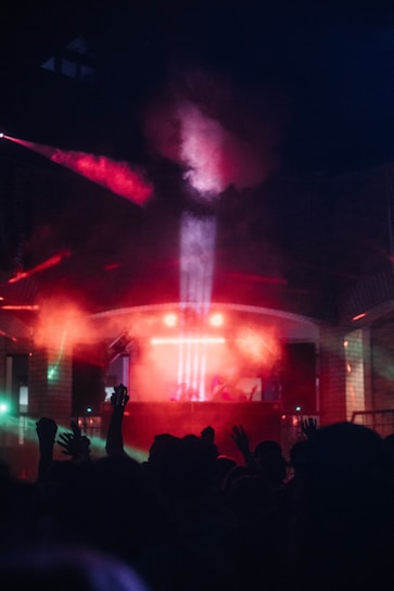 A vibrant club scene with colorful lighting effects, featuring a crowd of people with raised hands silhouetted against the bright lights. Smoky red and blue hues dominate the atmosphere as beams of light create a dynamic and energetic environment.
