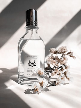 A glass perfume bottle with a shiny metallic cap stands next to delicate white flowers casting soft shadows on a light surface. The lighting creates elegant patterns, enhancing the minimalistic composition.