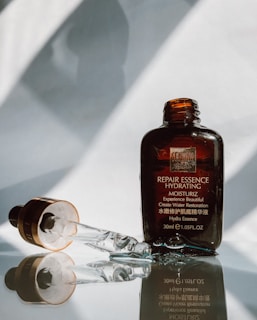 A translucent brown glass bottle labeled 'Repair Essence Hydrating' stands upright, accompanied by a dropper laying next to it. The background features soft shadows and reflections, creating a minimalistic and elegant presentation.