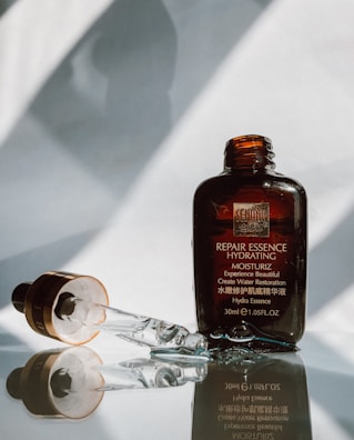 A translucent brown glass bottle labeled 'Repair Essence Hydrating' stands upright, accompanied by a dropper laying next to it. The background features soft shadows and reflections, creating a minimalistic and elegant presentation.